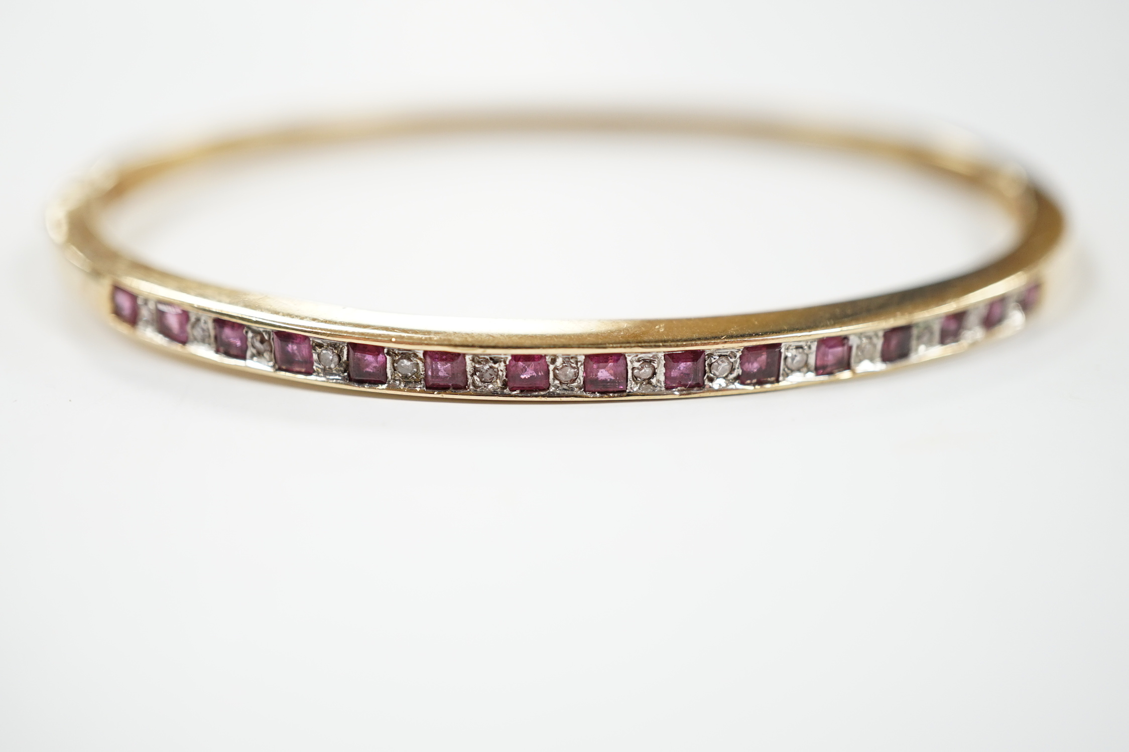 A yellow metal, ruby and diamond chip channel set hinged bangle, interior diameter 57mm, gross weight 10.8 grams.
